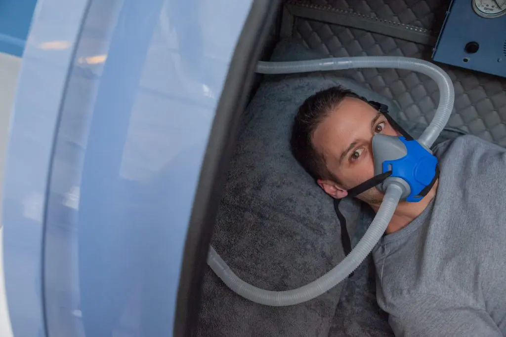 Person receiving hyperbaric oxygen therapy (HBOT) in a chamber while breathing through an oxygen mask.