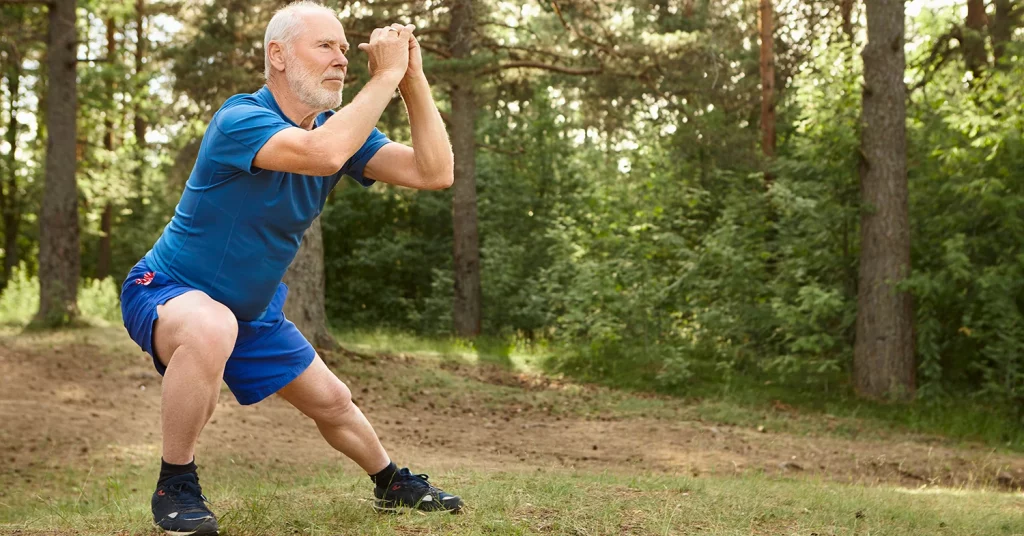 Portrait of healthy active elderly male pensioner in running shoes exercising outdoors, holding hands together in front of him and doing side lunges, having focused concentrated facial expression. Avoiding arthritis
