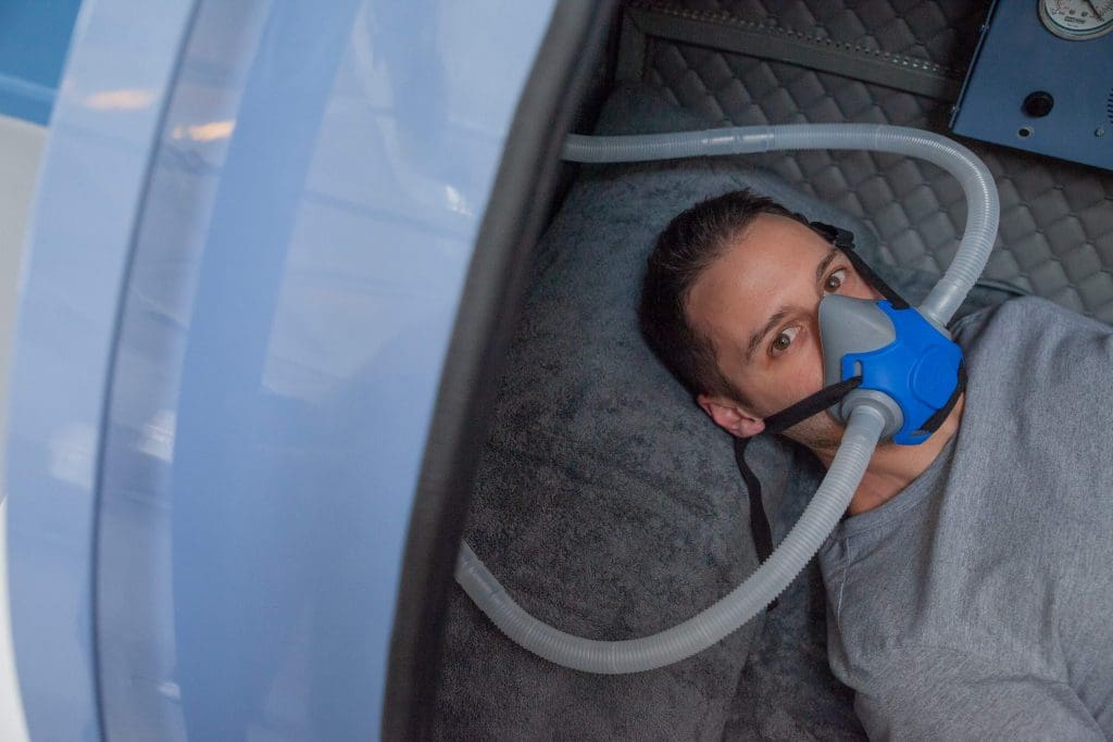 Person receiving hyperbaric oxygen therapy (HBOT) in a chamber while breathing through an oxygen mask.